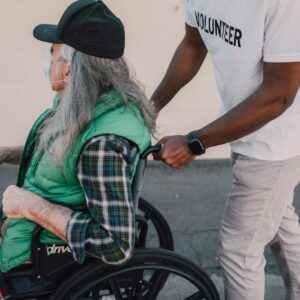 A volunteer helps a senior in a wheelchair during daytime outdoors.