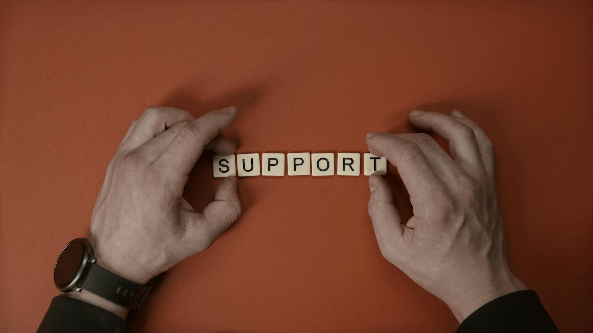 Hands arranging letter tiles to spell the word 'support' on a red background.